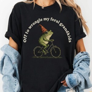 Op de afbeelding: Zwart T-shirt met een humoristische illustratie van een groene kikker met een puntmuts die op een fiets rijdt. De tekst "Off to wrangle my feral grandkids" staat boven de kikker. Het ontwerp is gedrukt op een zwart T-shirt.