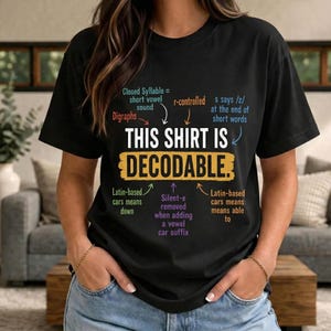 Op de afbeelding: Een zwart t-shirt met korte mouwen en de tekst THIS SHIRT IS DECODABLE in wit en geel. Rondom de tekst staan educatieve notities in verschillende kleuren, waaronder Closed Syllable, Digraphs, r-controlled en Latijnse definities.