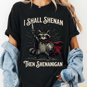 May include: Black t-shirt featuring a raccoon warrior with a sword and shield, wearing a red cape. The text on the shirt reads "I SHALL SHENAN THEN SHENANIGAN". The design is a humorous take on a classic hero.