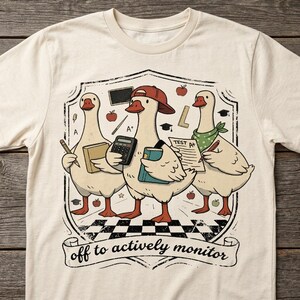 May include: Cream-colored t-shirt featuring a graphic of three geese dressed as students, each holding school supplies. The geese are inside a shield with apples and a banner that reads "off to actively monitor."