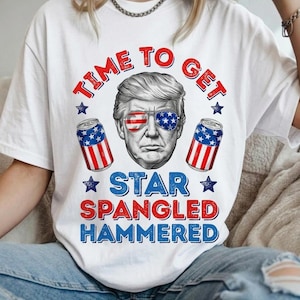 Puede incluir: Una camiseta blanca con un gráfico de un hombre que lleva gafas de sol con patrones de la bandera estadounidense. Dos latas de bebida con diseños de la bandera estadounidense flanquean el retrato. El texto dice: TIME TO GET STAR SPANGLED HAMMERED.