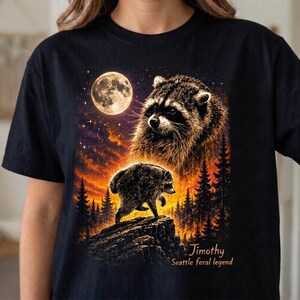 Puede incluir: Una camiseta negra con un estampado gráfico de la cara de un mapache grande y un mapache más pequeño sobre un acantilado rocoso. La escena incluye una luna llena, un cielo estrellado y una silueta de bosque con el texto Jimothy Seattle feral legend.