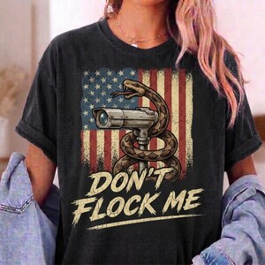 May include: A black short-sleeve t-shirt featuring a graphic of a coiled snake wrapped around a security camera, set against a distressed American flag. Below the graphic, the text reads DON'T FLOCK ME in a bold, weathered, light-coloured font.