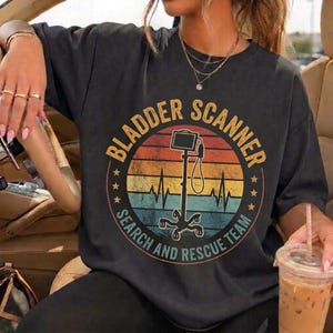 Puede incluir: Una camiseta gris carbón de manga corta con un gráfico circular sobre un fondo de atardecer retro. El diseño incluye una ilustración de un escáner de vejiga, una línea de latido cardíaco y el texto BLADDER SCANNER SEARCH AND RESCUE TEAM.