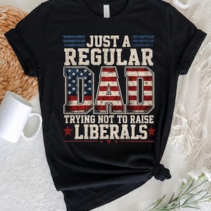 May include: Black t-shirt with the text "JUST A REGULAR DAD TRYING NOT TO RAISE LIBERALS" in a distressed, patriotic design. The word "DAD" is filled with an American flag pattern.