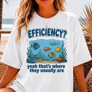 May include: A white short-sleeve t-shirt featuring a graphic of various colourful fish swimming in an underwater scene. The text EFFICIENCY? is printed above the fish, and the text yeah that's where they usually are is printed below.