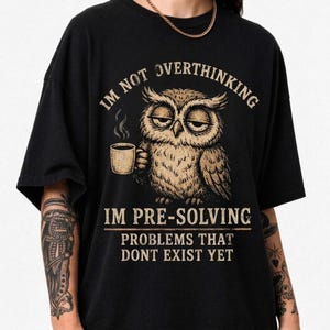 May include: A black t-shirt featuring a graphic of a tired-looking owl holding a steaming mug of coffee. The text reads: I'M NOT OVERTHINKING I'M PRE-SOLVING PROBLEMS THAT DONT EXIST YET. The design is rendered in shades of beige and brown.