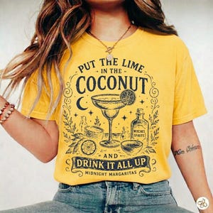 May include: A mustard yellow t-shirt featuring a black graphic print of a margarita glass, a bottle labelled Witches Spirits, and the text Put the Lime in the Coconut and Drink it All Up Midnight Margaritas with decorative stars and moon icons.