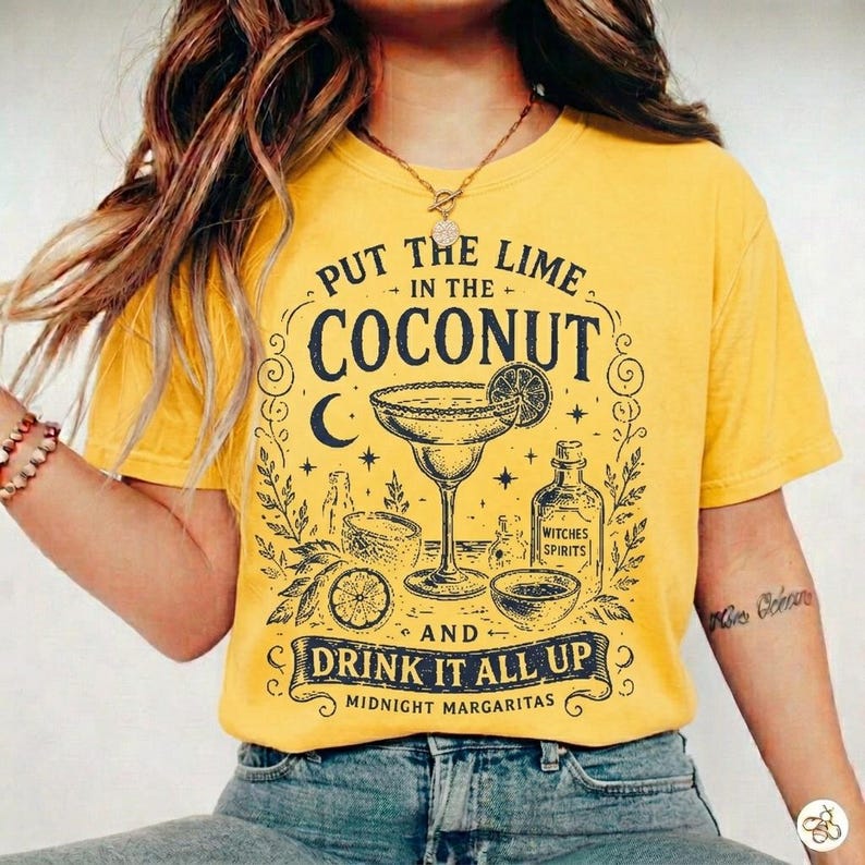 May include: A mustard yellow t-shirt featuring a black graphic print of a margarita glass, a bottle labelled Witches Spirits, and the text Put the Lime in the Coconut and Drink it All Up Midnight Margaritas with decorative stars and moon icons.