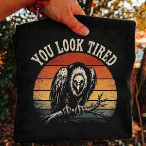 Può includere: Una t-shirt nera con la grafica di un avvoltoio appollaiato su un ramo davanti a un tramonto circolare multicolore. Il testo "YOU LOOK TIRED" è stampato in un carattere arcuato e dall'aspetto usurato sopra l'avvoltoio.