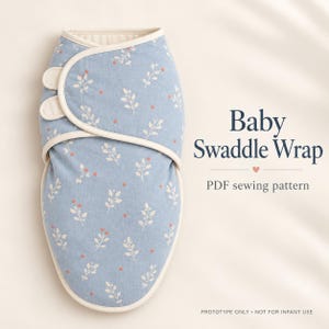 Könnte beinhalten: Ein hellblauer Baby-Pucksack mit weißem Blatt- und rotem Beerenmuster, cremefarbenen Rändern und zwei seitlichen Verschlüssen. Der Text lautet: Baby Swaddle Wrap, PDF sewing pattern, PROTOTYPE ONLY • NOT FOR INFANT USE.