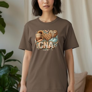 May include: A brown t-shirt featuring a colorful graphic design. The design includes a coffee cup, a sunflower, a heart with a leopard print, and the letters "CNA". The shirt is made of soft material.
