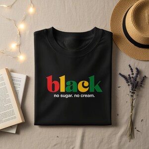 Może przedstawiać: Czarny t-shirt ze słowem "black" w kolorze czerwonym, żółtym i zielonym, z napisem "no sugar. no cream." poniżej. Koszulka jest złożona na beżowej powierzchni, z kapeluszem ze słomy i gałązką lawendy w tle.