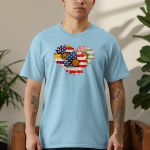 May include: Light blue t-shirt featuring a heart-shaped floral design with American flag elements. The design incorporates red, white, and blue colors, resembling the American flag. The shirt is made of a soft material and has short sleeves.