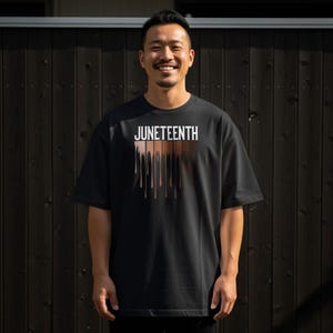 Könnte beinhalten: Schwarzes T-Shirt mit dem weißen Wort "JUNETEENTH". Darunter befinden sich vertikale Linien in verschiedenen Brauntönen, die Hauttöne darstellen. Die Person, die das T-Shirt trägt, lächelt.
