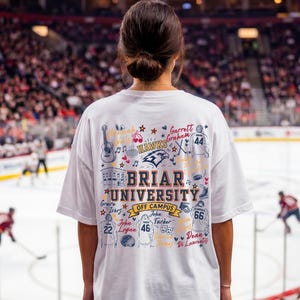 May include: A white t-shirt featuring a colorful graphic on the back with the text Briar University Off Campus, surrounded by illustrations of hockey jerseys, a guitar, a building, and various names written in cursive script and block lettering.