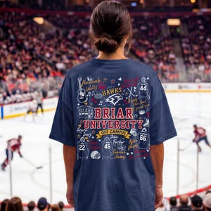 May include: A navy blue t-shirt featuring a graphic print on the back with the text Briar University Off Campus, surrounded by illustrations of hockey jerseys, a guitar, a building, and various names written in a decorative script font.