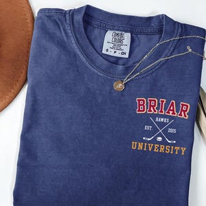 May include: A navy blue short-sleeve t-shirt with a crew neckline. The chest features a graphic with the text BRIAR UNIVERSITY in red and yellow, and HAWKS EST. 2015 with crossed hockey sticks and a puck. A gold-toned necklace rests on the shirt.