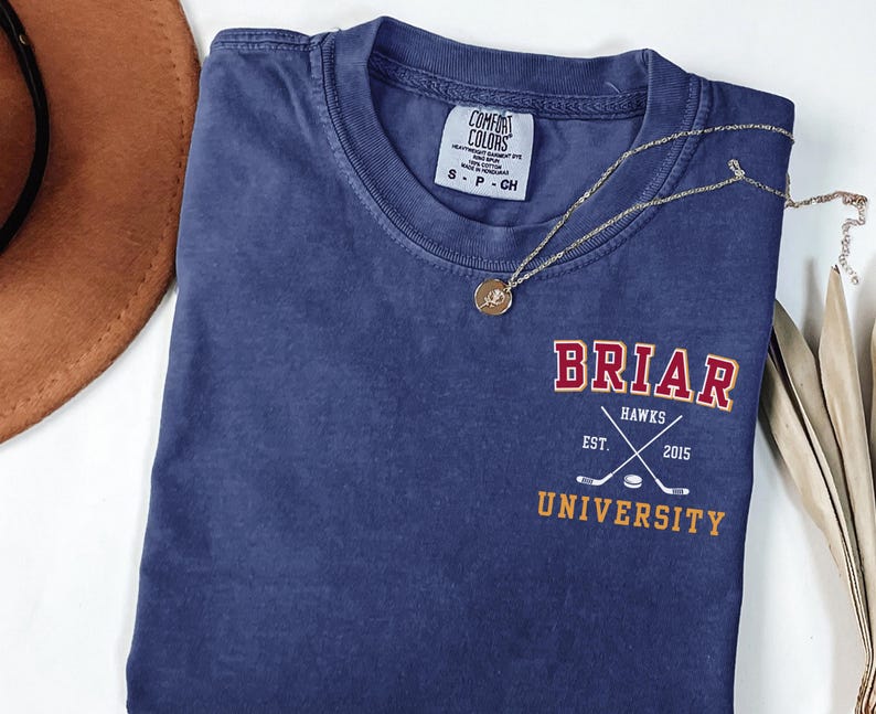 May include: A navy blue short-sleeve t-shirt with a crew neckline. The chest features a graphic with the text BRIAR UNIVERSITY in red and yellow, and HAWKS EST. 2015 with crossed hockey sticks and a puck. A gold-toned necklace rests on the shirt.