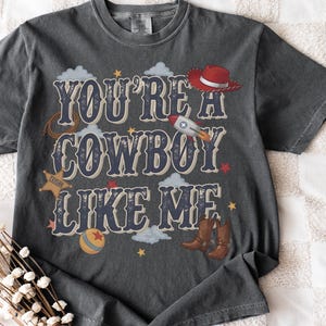 May include: A charcoal gray short-sleeve t-shirt featuring the text "YOU'RE A COWBOY LIKE ME" in a bold, distressed font. The graphic includes a red cowboy hat, a rocket ship, a sheriff badge, a lasso, a ball, and brown cowboy boots.