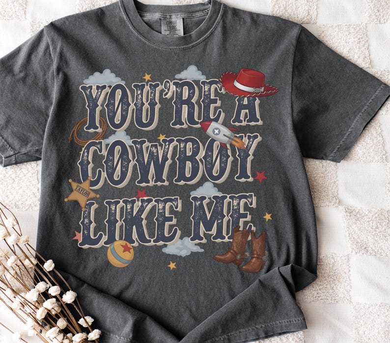 May include: A charcoal gray short-sleeve t-shirt featuring the text "YOU'RE A COWBOY LIKE ME" in a bold, distressed font. The graphic includes a red cowboy hat, a rocket ship, a sheriff badge, a lasso, a ball, and brown cowboy boots.