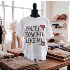 May include: A white short-sleeve t-shirt featuring the text "YOU'RE A COWBOY LIKE ME" in a dark blue font. The graphic includes illustrations of a red cowboy hat, a rocket ship, a lasso, a yellow star, a ball, and brown cowboy boots.