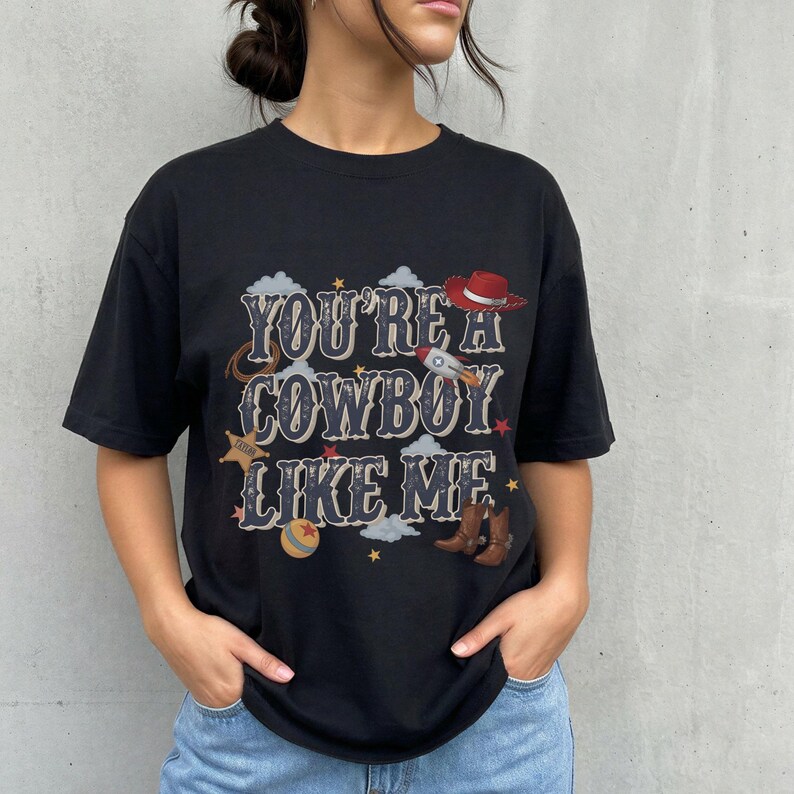 May include: A black short-sleeved t-shirt featuring the text "YOU'RE A COWBOY LIKE ME" in a decorative font. The graphic includes illustrations of a red cowboy hat, a rocket, a sheriff star, a lasso, a ball, and brown cowboy boots.