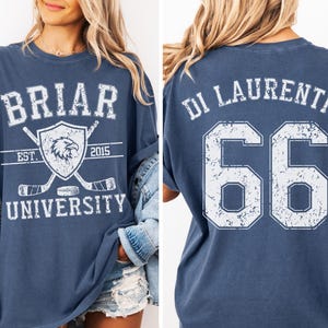 Könnte beinhalten: Ein blaues Kurzarm-T-Shirt mit einem abgenutzten weißen Aufdruck. Die Vorderseite zeigt den Text BRIAR UNIVERSITY, ein Adler-Emblem, Hockeyschläger und EST. 2015. Die Rückseite zeigt den Text DI LAURENTIS und die große Zahl 66 in Weiß.