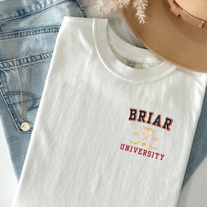May include: A white short-sleeve t-shirt featuring a graphic on the left chest that reads BRIAR UNIVERSITY in navy and red collegiate lettering. The design includes crossed hockey sticks and a puck in yellow with the text HAWKS EST 2015.