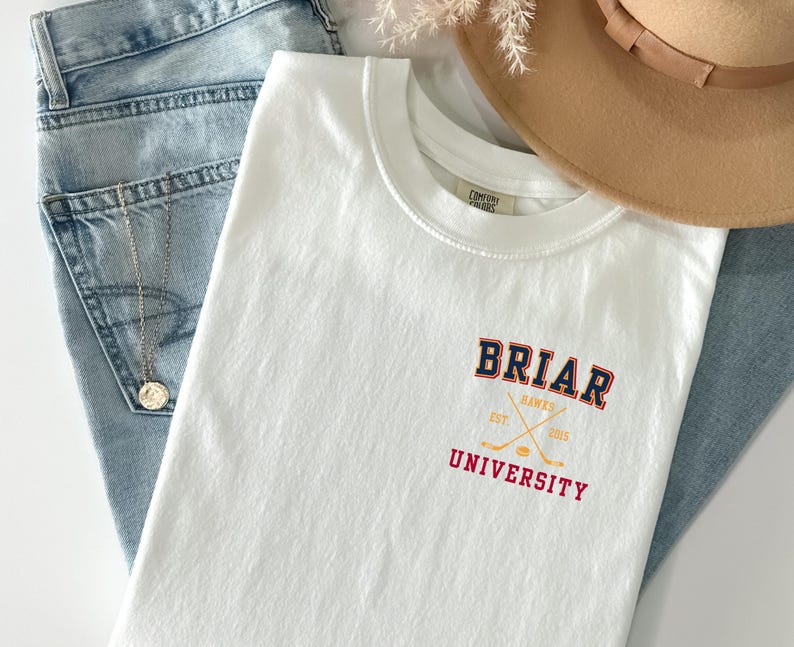 May include: A white short-sleeve t-shirt featuring a graphic on the left chest that reads BRIAR UNIVERSITY in navy and red collegiate lettering. The design includes crossed hockey sticks and a puck in yellow with the text HAWKS EST 2015.