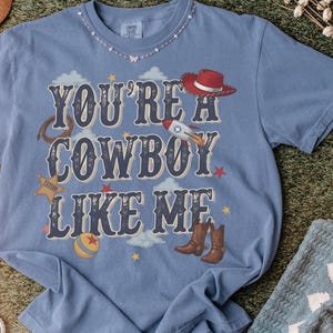 May include: A blue short-sleeve t-shirt featuring the text "YOU'RE A COWBOY LIKE ME" in dark blue lettering. The design includes illustrations of a red cowboy hat, a rocket, a lasso, a sheriff star, a ball, and brown cowboy boots among clouds.