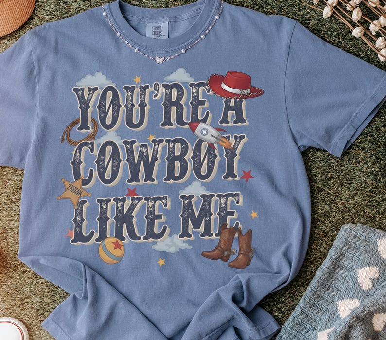 May include: A blue short-sleeve t-shirt featuring the text "YOU'RE A COWBOY LIKE ME" in dark blue lettering. The design includes illustrations of a red cowboy hat, a rocket, a lasso, a sheriff star, a ball, and brown cowboy boots among clouds.