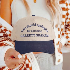 May include: A beige baseball cap with a dark navy blue brim and dark navy blue embroidered text that reads: men should apologize for not being GARRETT GRAHAM. The cap features a structured crown and a button at the top center.