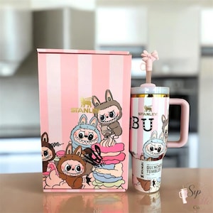 May include: A pink and white striped Stanley Quencher tumbler with a matching box, featuring cartoon characters and a bow-shaped straw topper. The tumbler has a handle, a gold-colored rim, and text that reads Stanley and Quencher Tumbler.