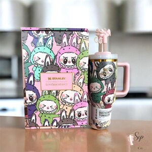 May include: A Stanley LoveShackFancy Quencher tumbler with a matching box, both featuring a repeating pattern of cartoon characters in bunny suits. The tumbler has a pink handle, a gold rim, and a pink bow-shaped straw topper on a clear straw.