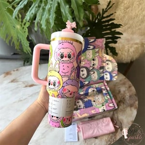 May include: A pink tumbler with a handle and a lid featuring a bow, decorated with a repeating pattern of cartoon creatures in various colors. The tumbler has a white label with black text and is positioned in front of a matching patterned box.