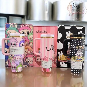 May include: Three stainless steel tumblers with handles and lids, each featuring Labubu character designs. One is pink with a multi-colored character pattern, one is pink with vertical stripes and Labubu text, and one is black with white patterns.
