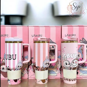 May include: Three pink and white striped tumblers with handles and lids, each featuring Labubu character graphics and text. The tumblers are positioned in front of matching pink striped boxes labeled Labubu, with a Sip & Sparkle Co. logo visible.