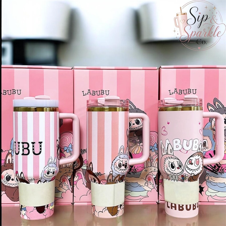 May include: Three pink and white striped tumblers with handles and lids, each featuring Labubu character graphics and text. The tumblers are positioned in front of matching pink striped boxes labeled Labubu, with a Sip & Sparkle Co. logo visible.