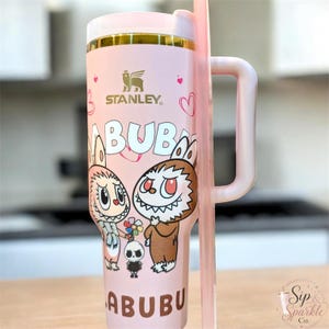 May include: A light pink Stanley tumbler with a handle and straw, featuring a gold rim and a graphic print of two Labubu characters with sharp teeth, a small skull figure, and hearts. The text "LABUBU" is printed in brown at the top and bottom.