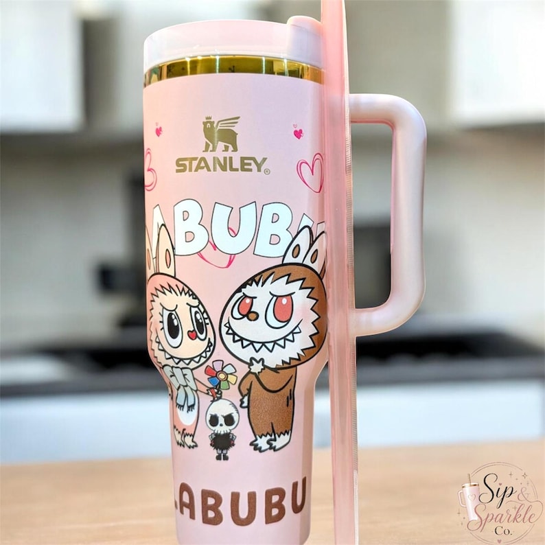 May include: A light pink Stanley tumbler with a handle and straw, featuring a gold rim and a graphic print of two Labubu characters with sharp teeth, a small skull figure, and hearts. The text "LABUBU" is printed in brown at the top and bottom.