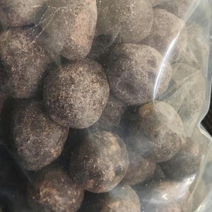 Puede incluir: Una bolsa de plástico transparente llena de numerosos caramelos redondos y texturizados de color marrón oscuro. Los caramelos tienen una superficie rugosa y desigual y parecen estar cubiertos de un polvo fino. La impresión general es la de un artículo a granel.