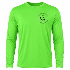 Op de afbeelding: Een fel neongroen shirt met lange mouwen en een zwart cirkelvormig logo op de linkerborst. Het logo bevat de letters CA in het midden met de tekst CUSTOM GIFTS CREATIONSAMOR in een cirkel rondom de letters gerangschikt.