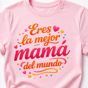 May include: A light pink t-shirt with the Spanish text "Eres la mejor mamá del mundo" in orange and red lettering. The design includes hearts and dots in various colors.