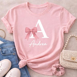 Op de afbeelding: Roze t-shirt met de tekst "Personalized Name Shirt" en een hartje. Het shirt heeft een grote witte letter "A" met een roze strik en de naam "Andrea" in witte letters. Het wordt gestyled met een jeans, witte sneakers en een handtas met parels.
