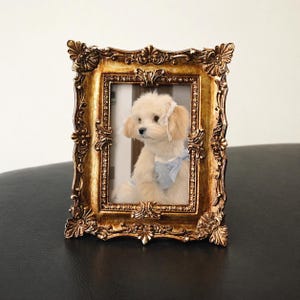 May include: A small, fluffy dog in a blue and white outfit and a white headpiece is framed in an ornate, gold-coloured picture frame. The frame has detailed scrollwork and a vintage aesthetic, adding a touch of elegance to the image.
