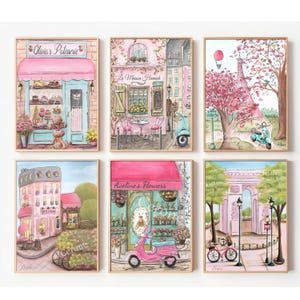 Op de afbeelding: Zes ingelijste aquarel schilderijen tonen charmante Parijse sc&egrave;nes. Elk toont een winkel of herkenningspunt, zoals een patisserie, bloemenwinkel en de Eiffeltoren, in zachte roze, blauwe en groene tinten. Scooters en fietsen dragen bij aan de grillige, romantische sfeer.
