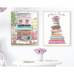 May include: Two framed watercolor prints. One depicts a pink building labeled "Olivia's Bookshop" with a blue awning. The other shows a stack of books topped with flowers, with the text "Olivia's Books" above. Both are in gold frames.