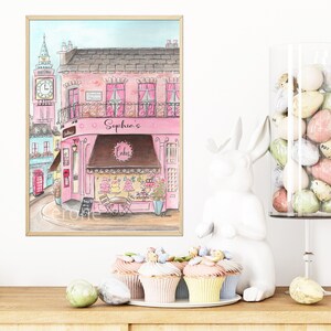 May include: A watercolor painting of a pink building with a sign that says "Sophia's Cakes". The building is on a street with a red telephone booth and a clock tower. There are cupcakes on a white plate in front of the painting.