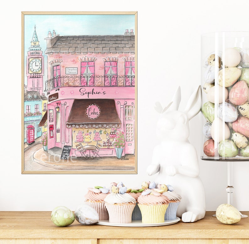 May include: A watercolor painting of a pink building with a sign that says "Sophia's Cakes". The building is on a street with a red telephone booth and a clock tower. There are cupcakes on a white plate in front of the painting.